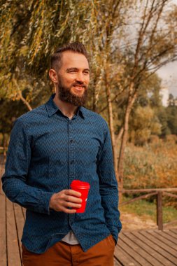 Güz parkında sabah kahvesi içip yürüyen orta yaşlı, yakışıklı, sakallı adam..