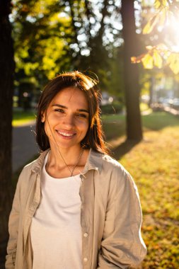 Gün batımında gülümseyen ve parka bakan güzel Amerikalı kadın portresi. Gülümseyen bir kızın açık hava portresi. Mutlu neşeli kız parka gülüyor. 