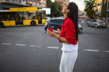 Sokak geçidinde arka planda akıllı telefon ve kahve tutan yakın plan kadın elleri. Şehir yolunda duran mobil cihaz kullanan genç iş kadını