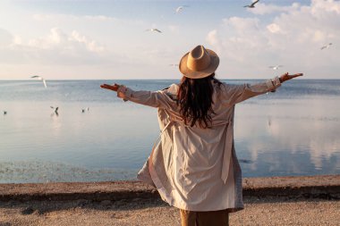 Young happy asian woman with hands in the air walks on the seaside in autumn. Seagulls flying on the beach.