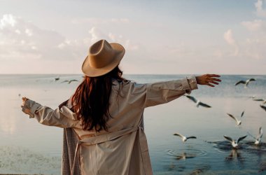 Young happy asian woman with hands in the air walks on the seaside in autumn. Seagulls flying on the beach.
