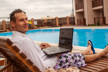 Businessman lying in chaise-lounge by swimming pool and checking e-mails on laptop. Positive man using laptop on summer vacation.
