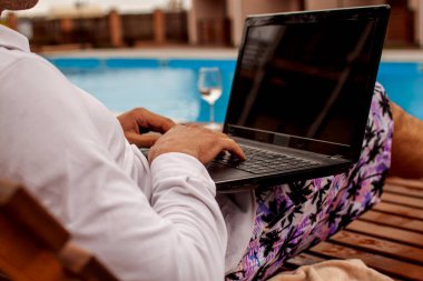 Man lying in chaise-lounge by swimming pool and checking e-mails on laptop. Positive man using laptop on summer vacation.