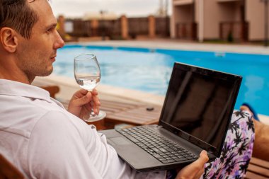 Businessman lying in chaise-lounge by swimming pool and checking e-mails on laptop. Positive man using laptop on summer vacation.