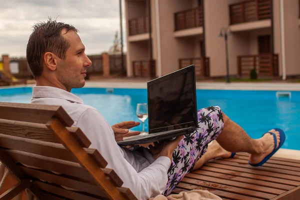 Businessman lying in chaise-lounge by swimming pool and checking e-mails on laptop. Positive man using laptop on summer vacation.