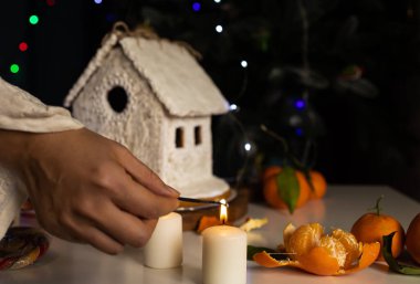 A small gingerbread house with frosting, standing on a table with decorations, candles and tangerines. Festive mood
