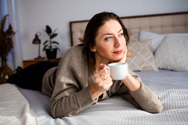 Mutlu, orta yaşlı bir kadın elinde bir fincan çay ya da kahveyle sabah yatak odasında sıcak bir gülümsemeyle..