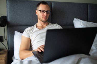 Tişört giyen, gülümseyen genç sakallı adam dizüstü bilgisayarla çalışıyor çizgili yastık battaniyesiyle yatakta yatıyor yatak odasında dinleniyor.