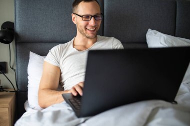 Tişört giyen, gülümseyen genç sakallı adam dizüstü bilgisayarla çalışıyor çizgili yastık battaniyesiyle yatakta yatıyor yatak odasında dinleniyor.