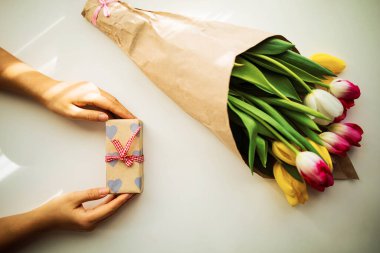 Bir kutu dolusu lalenin yanında hediye taşıyan kadın eller, üst manzara.
