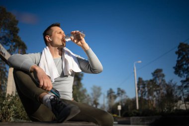 Bir şişe suyla parkta dinlenen bir sporcu, cep telefonuyla sargı bezi ve müzik dinleyen (kasıtlı güneş parıltısı ve klasik renk))