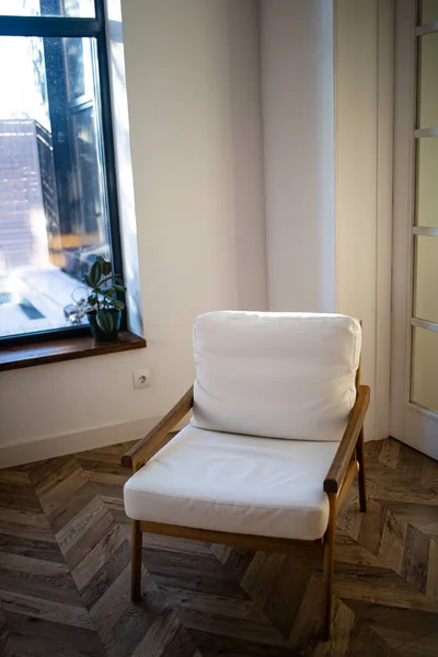 White soft armchair next to window in living room interior with old wooden floor.