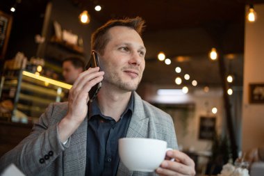 Genç iş adamı kahve dükkanında oturmuş akıllı telefonla konuşuyor.