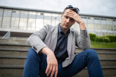 Stresli, takım elbiseli, elleri başının üstünde ofis binasının yanındaki merdivenlerde oturan genç bir iş adamı. İşsizlik ve işten çıkarma kavramı.