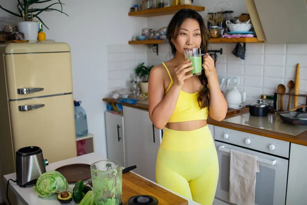 Fit girl preparing green detox juice with spinach and celery at home after training, wearing sport clothes