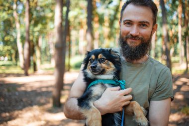 Yakışıklı sakallı adam parkta köpeğini kucaklıyor..