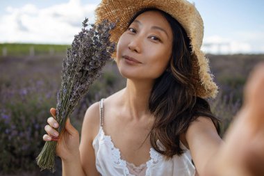 Hasır şapkalı mutlu Asyalı genç kız tarlada lavanta buketi tutuyor, selfie çekiyor..