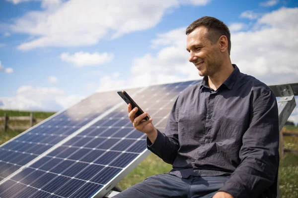 a man holds a smartphone near solar panels on a plot near the house.