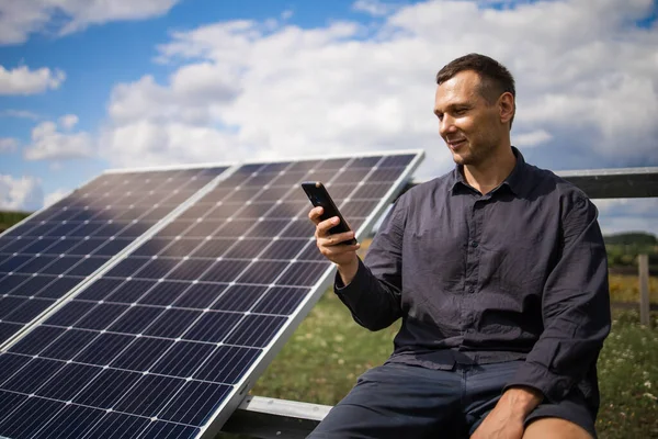 a man holds a smartphone near solar panels on a plot near the house.
