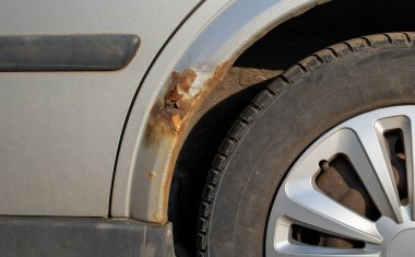 Part of a silver car with rust and a holes in the wing and a black tire with a deep off-road tread