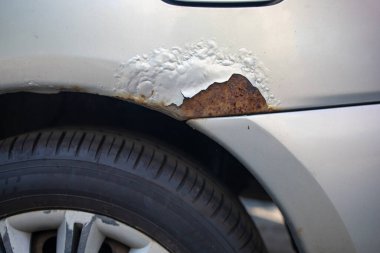 Part of a silver car with rust and a holes in the wing and a black tire with a deep off-road tread