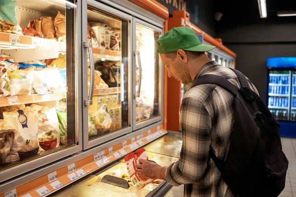 Young man chooses frozen food products at grocery supermarket. Young man shopping in supermarket, reading product information.
