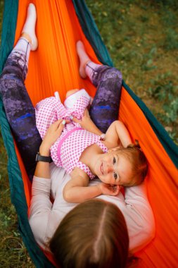 young mother and cute little daughter relaxing together and smiling sitting in a hammock, hugging and lounging during a sunny summer day in a holiday home garden with grass and trees, lifestyle.