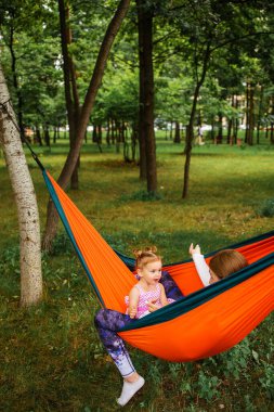 young mother and cute little daughter relaxing together and smiling sitting in a hammock, hugging and lounging during a sunny summer day in a holiday home garden with grass and trees, lifestyle.