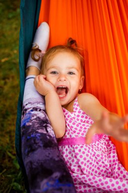 young mother and cute little daughter relaxing together and smiling sitting in a hammock, hugging and lounging during a sunny summer day in a holiday home garden with grass and trees, lifestyle.