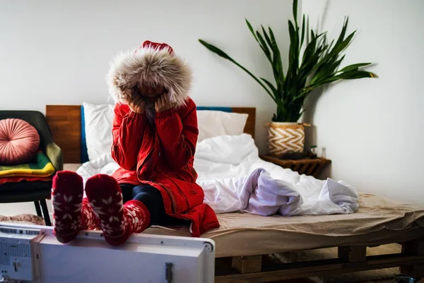 Young cold woman in a winter jacket sitting next to an electric heater at home.