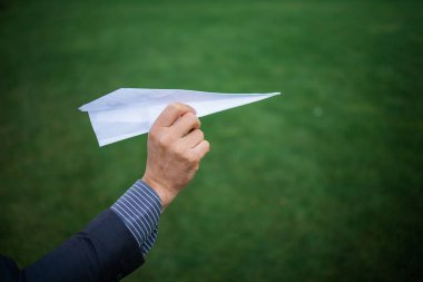 Businessman's hand preparing to throwing paper plane to mid air against green grass field with copy space. 