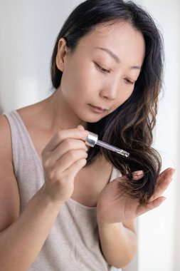 Ayrılmak Bakımı bitirir. Gülümseyen Asyalı Kadın kıvırcık saçlarına nemlendirici yağ sürerken genç bir kadının evde güzellik rutini yaparken saç bakım ürünlerini damlatırken kapatması..