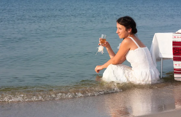 Молодая девушка в мокром белом платье сидит с бокалом шампанского в руке у моря — стоковое фото