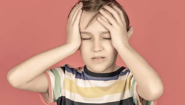 Huvudvärk barn. Lider av migrän. Huvudvärk eftersom stress. Porträtt av en ledsen pojke som håller huvudet med handen, isolerad på den röda bakgrunden. En liten pojke med huvudvärk. förtvivlan, tragedi — Stockfoto