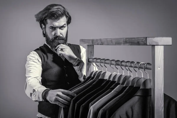 Roupas de homem, boutiques. Alfaiate, alfaiataria. Fato de homem elegante. Fato de homem, alfaiate na oficina. Bonito barbudo homem de moda em traje clássico. ternos masculinos pendurados em uma fileira — Fotografia de Stock