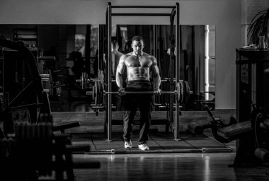Spor salonunda halterle çalışan kaslı bir adam. Vücut geliştirici atletik adam, karın kasları, omuzları, pazıları, kol ve göğsü mükemmel. Koltuk değneği egzersizi. Siyah ve beyaz