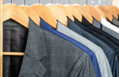 Perakende giyim mağazasında askıda asılı farklı renklerde erkek giysileri, yakın plan. Erkek gömlekleri, takım elbise askıda asılı. Butikte üzerinde ceket olan askılar. Askıda asılı erkekler için takım elbiseler