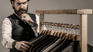Erkek kıyafetleri, butikler. Terzi, terzilik. Şık erkek kıyafeti. Erkek kıyafeti, atölyesinde terzi. Klasik kostüm giymiş yakışıklı sakallı moda adamı. Erkek takımları peş peşe asılıyor