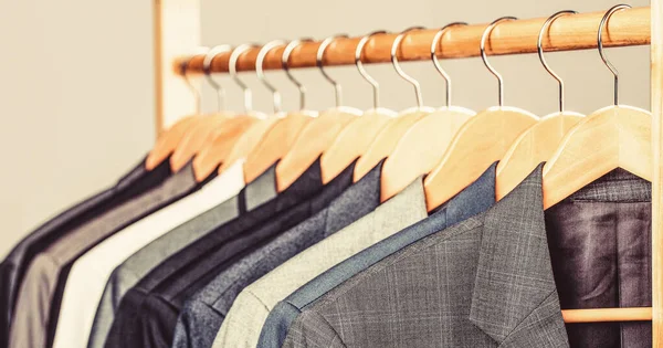 Trajes para hombre en diferentes colores que cuelgan de la percha en una tienda de ropa al por menor, primer plano. Camisas para hombre, traje colgado en el estante. Perchas con chaquetas en ellos en boutique. Trajes para hombres colgados en el bastidor — Foto de Stock