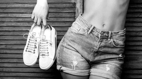 Mulher segurando sapatos em um fundo azul. Mulher segurando um par de sapatos brancos. A rapariga tem um par de sapatos. Menina com uma cintura bonita em calções de ganga. Mulher em um fundo azul com uma barriga plana — Fotografia de Stock