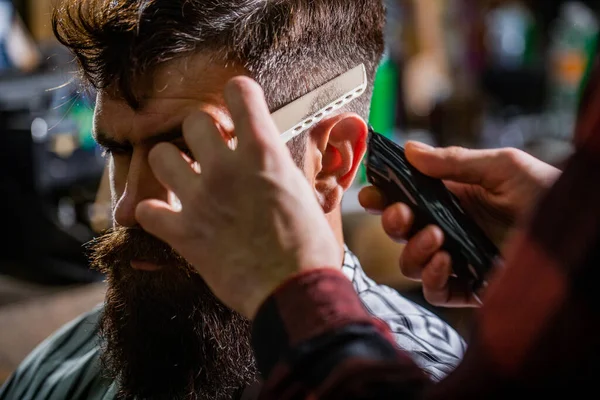 Conceito de corte de cabelo. Homem visitando cabeleireiro na barbearia. Barbeiro trabalha com cortador de cabelo. Um cliente Hipster a cortar o cabelo. Mãos de barbeiro com cortador de cabelo, close-up — Fotografia de Stock