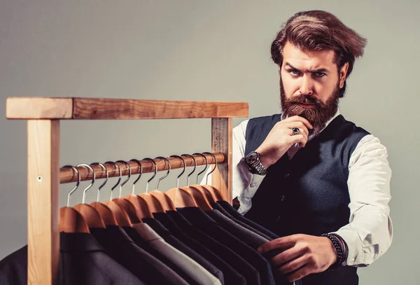 Men clothing, boutiques. Tailor, tailoring. Stylish mens suit. Man suit, tailor in his workshop. Handsome bearded fashion man in classical costume suit. Male suits hanging in a row