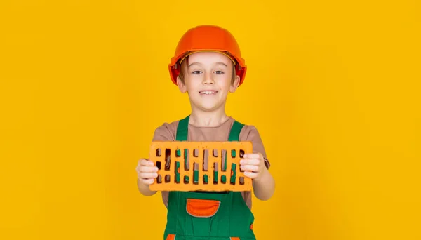Boy in a construction helmet holds a brick in his hands on yellow background. Little builder in helmet. Child dressed as a workman builder. Little boy wearing helmet. Child building helmet — Stock Photo, Image