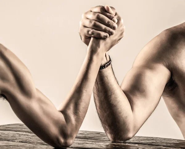 Arms wrestling thin hand, big strong arm in studio. Two mans hands clasped arm wrestling, strong and weak, unequal match. Arm wrestling. Heavily muscled bearded man arm wrestling a puny weak man Stock Picture