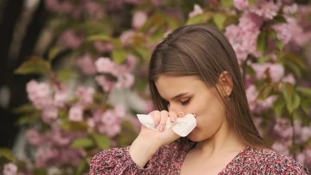 Spring allergy concept. Sneezing young girl with nose wiper among blooming trees in park. Pollen allergy, girl sneezing — Vídeo de stock