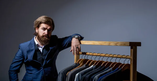 Stylish mens suit. workshop. Handsome bearded fashion man in classical costume suit. Male suits hanging in a row. Men clothing, boutiques. Man suit, tailor in his. Tailor, tailoring.