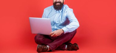 Gülümseyen yakışıklı sakallı işçi dizüstü bilgisayar. Kırmızı arka planda dizüstü bilgisayarla yerde oturan mutlu genç adam..