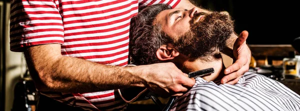 Barber shaving a bearded man in a barber shop. Closeup. Men barber shaves his beard. Bearded male sitting in an armchair in a barber shop while hairdresser shaves his beard with a dangerous razor.