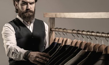 Stylish mens suit. Handsome bearded fashion man in classical costume suit. Man suit, tailor in his workshop. Male suits hanging in a row. Tailor, tailoring.