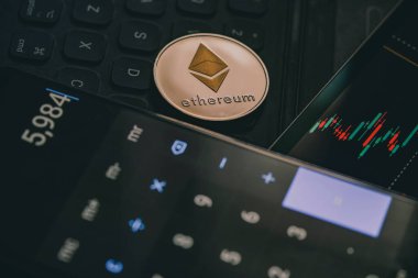 Close up of an Ethereum cryptocurrency coin near a monitor with candle stick graph chart and digital calculator.